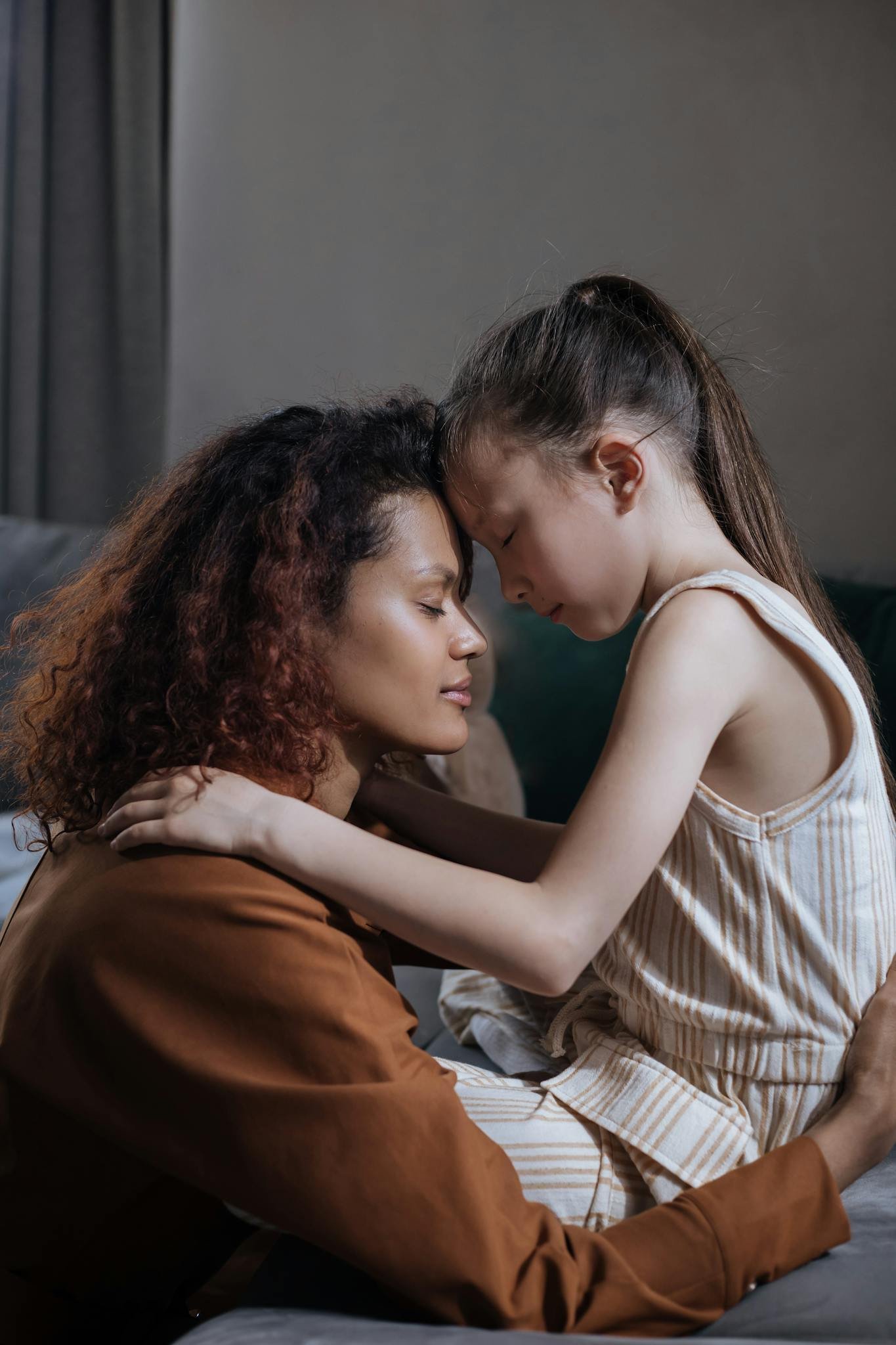 A mother and daughter share a tender moment indoors, embracing with eyes closed.