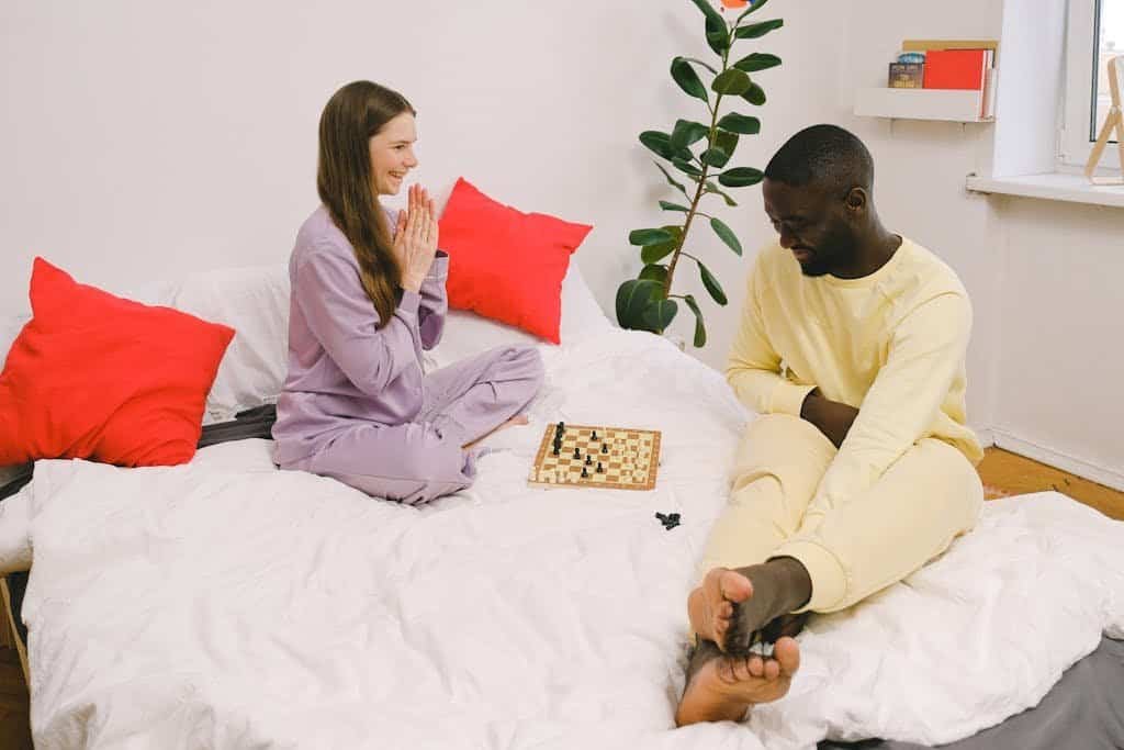 A happy interracial couple in pajamas plays chess on a bed, bonding and smiling.